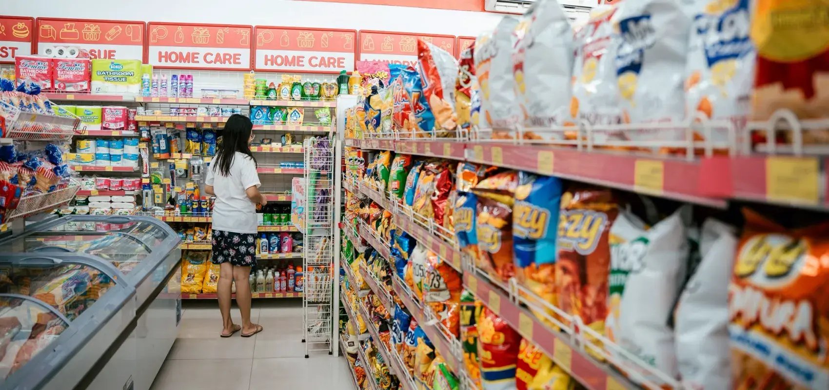 Convenience store aisle with shelves of snacks, household goods, and freezers